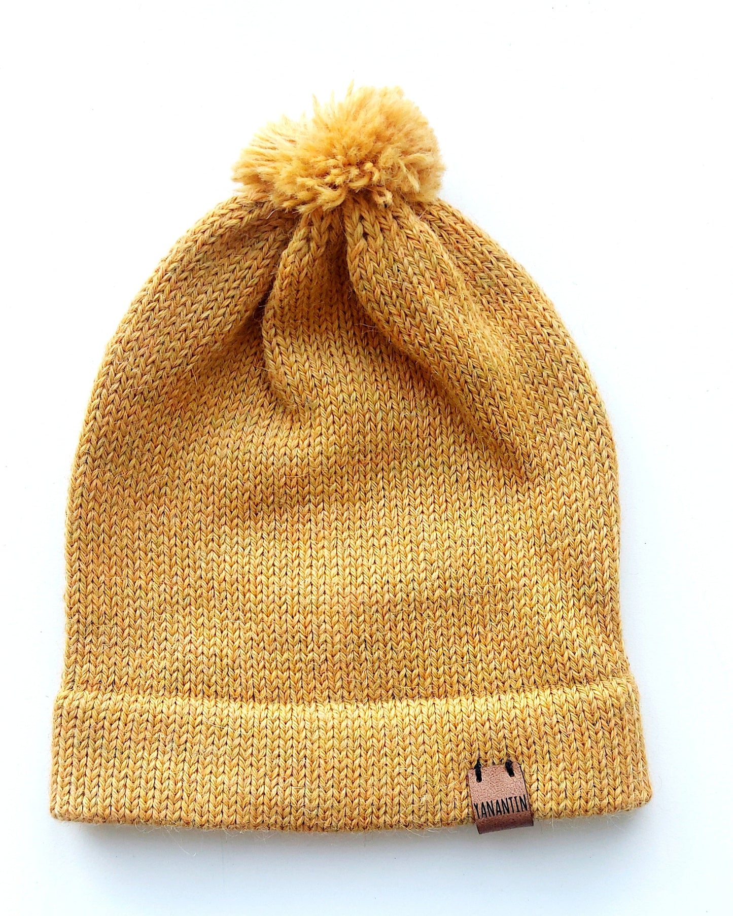 Handmade light yellow baby hat in soft 100% baby alpaca wool with a pompom, foldable edge, and brown vegan leather label, offering cozy warmth and cute design.