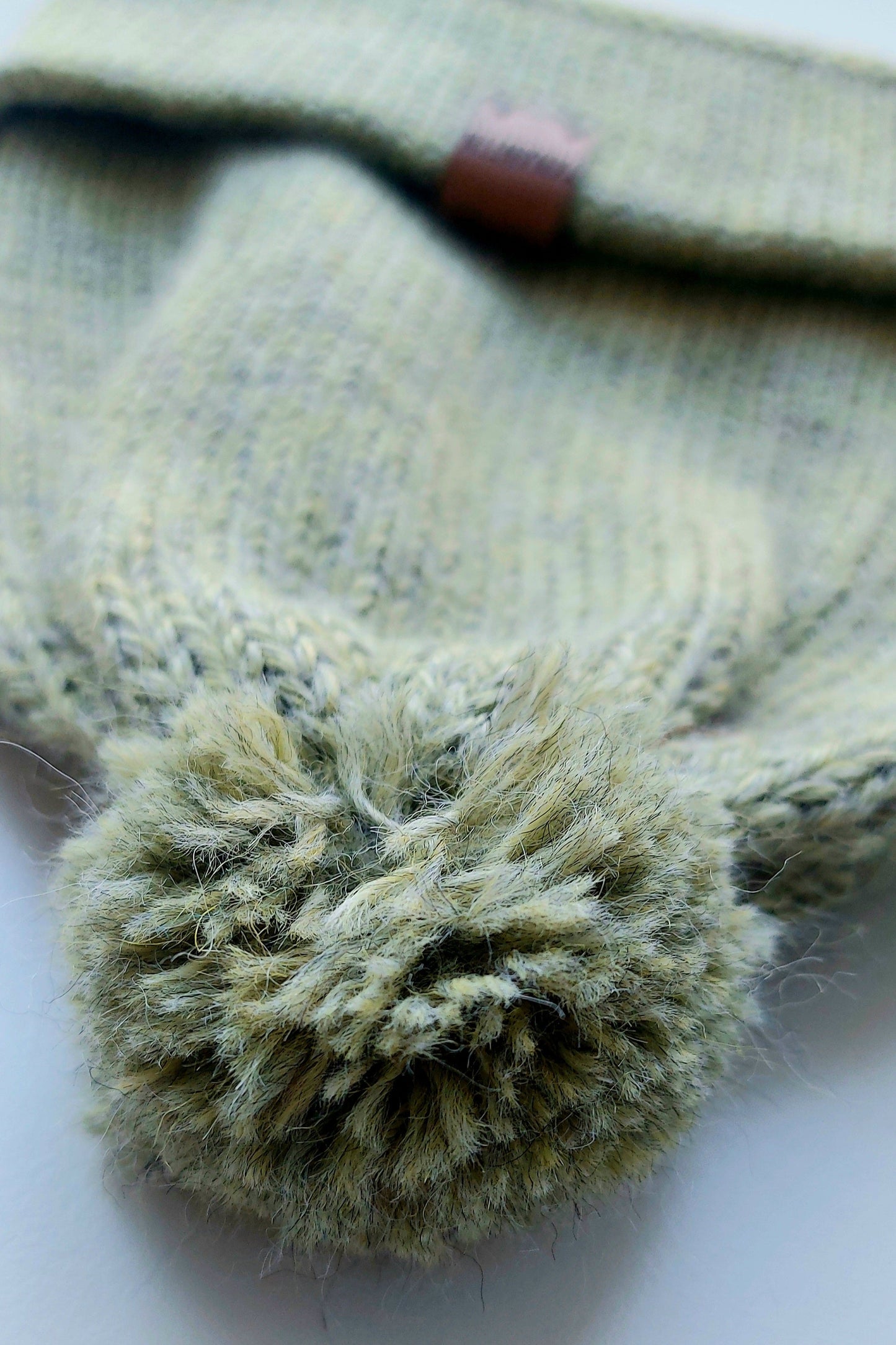 Close-up of the pompom on the handmade light green baby hat, made from soft 100% baby alpaca wool with a foldable edge and brown vegan leather label.