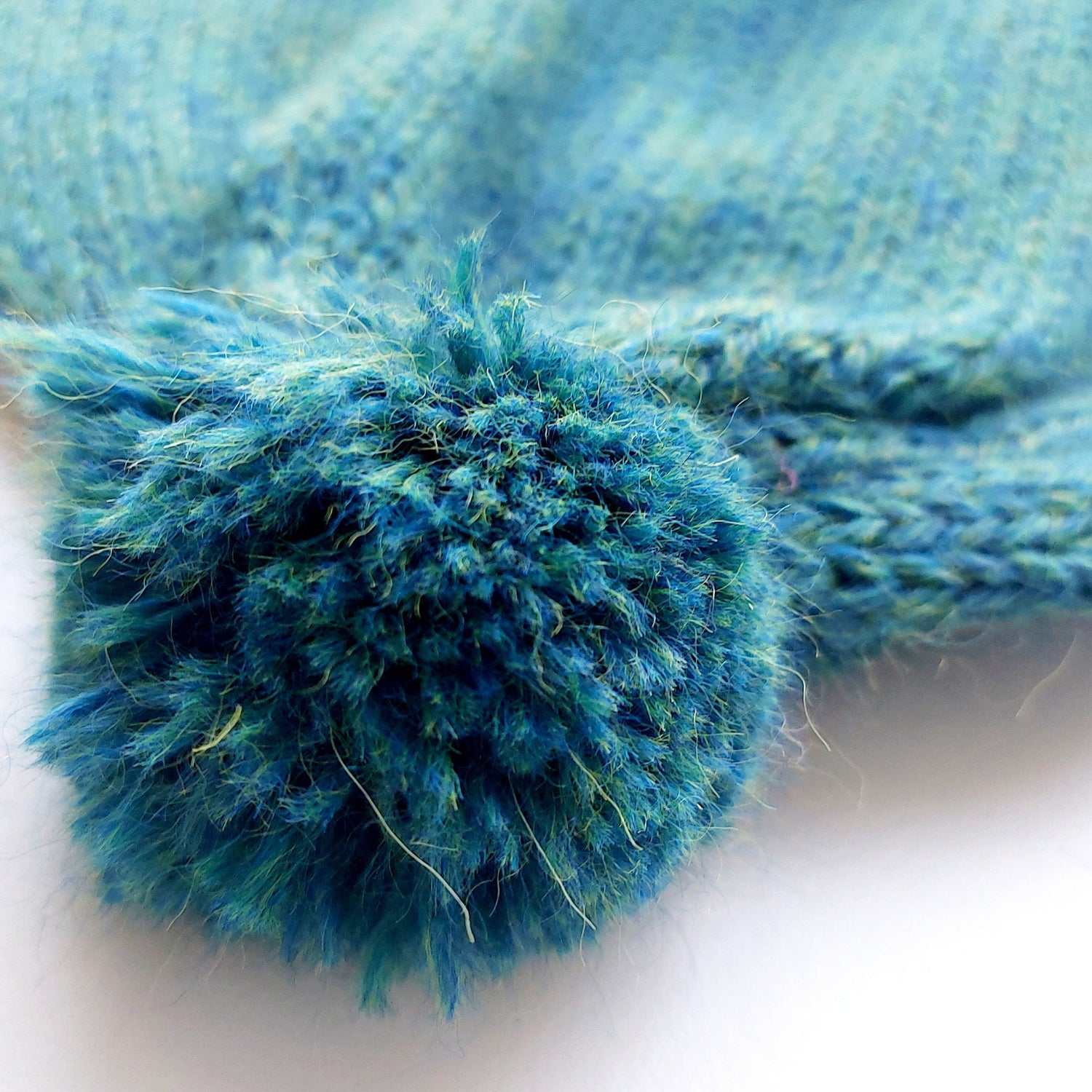 Close-up of the pompom on the handmade peacock blue baby hat, made from soft 100% baby alpaca wool with a foldable edge and brown vegan leather label.