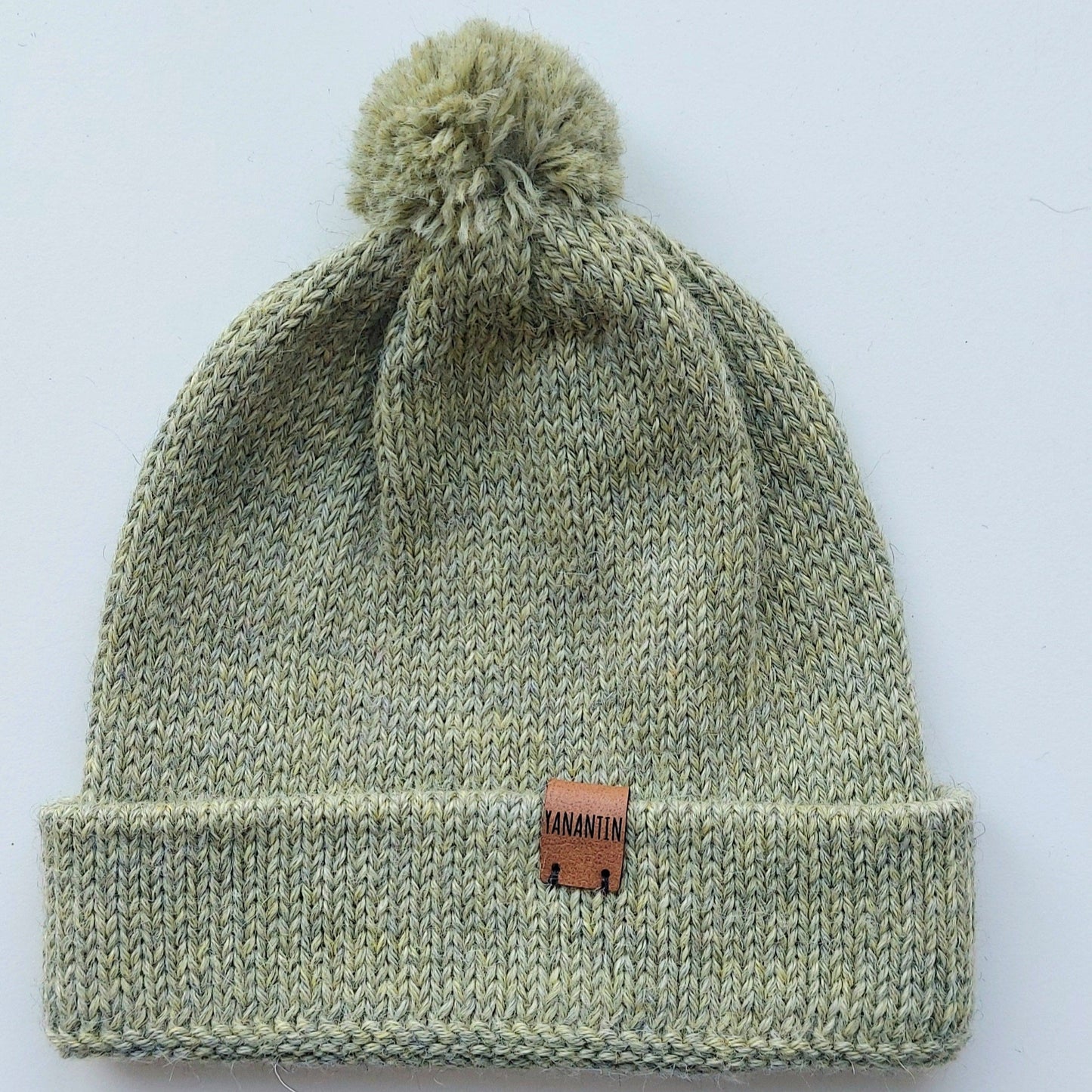 Handmade light green baby hat in soft 100% baby alpaca wool with a pompom, foldable edge, and brown vegan leather label, offering warmth and style.