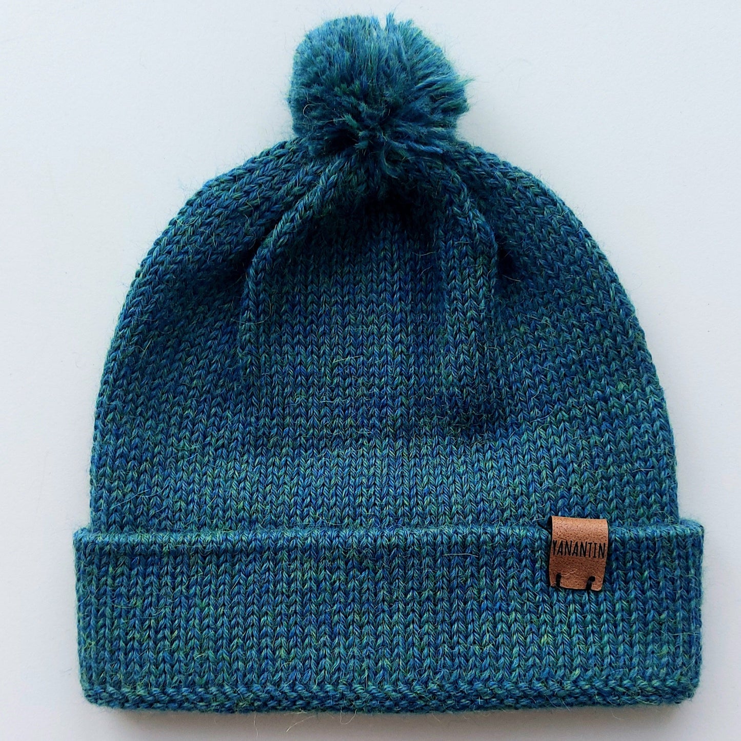 Handmade peacock blue baby hat in soft 100% baby alpaca wool with a pompom, foldable edge, and brown vegan leather label, offering warmth and comfort.