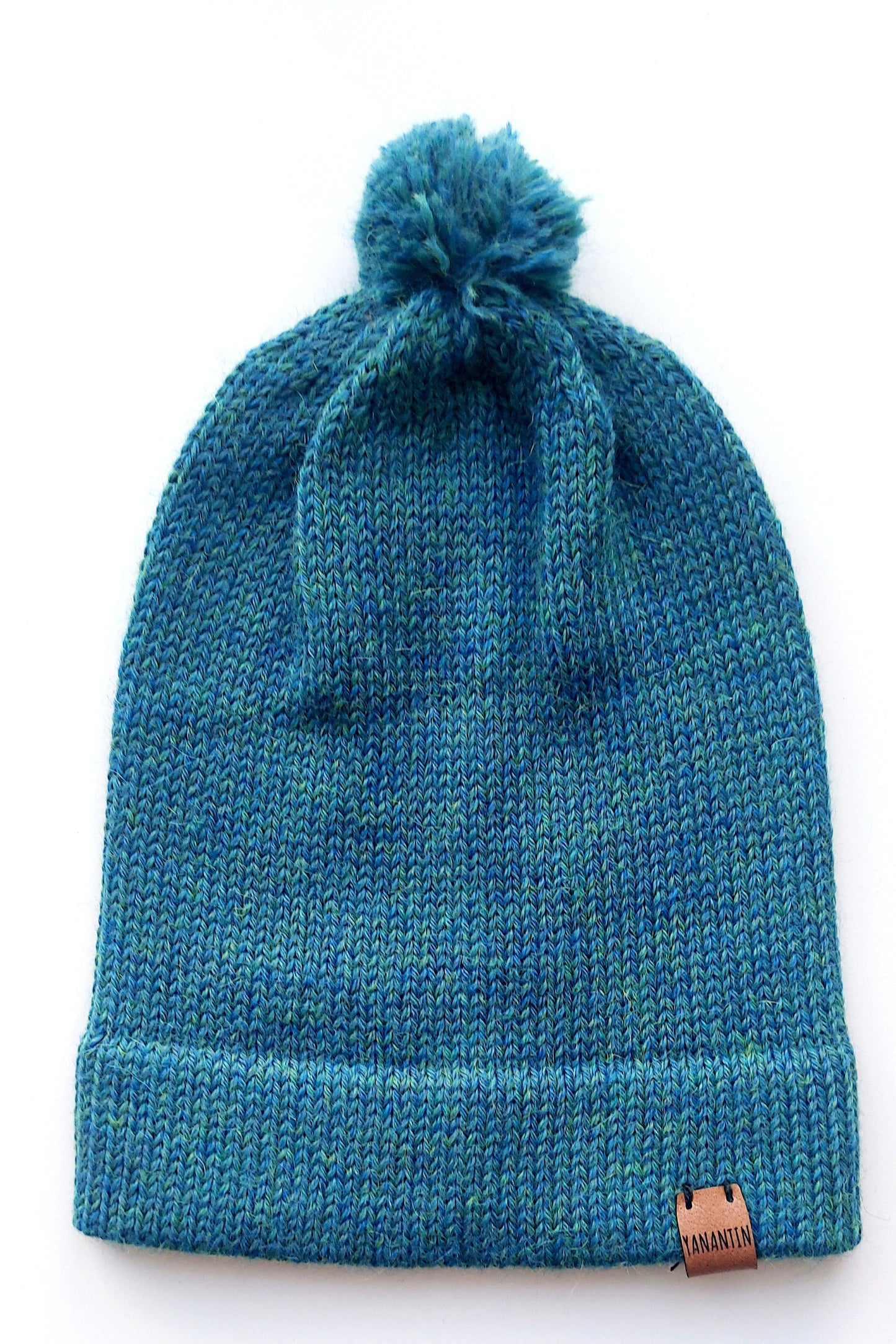 Handmade peacock blue baby hat in soft 100% baby alpaca wool with a pompom, foldable edge, and brown vegan leather label, offering warmth and comfort.