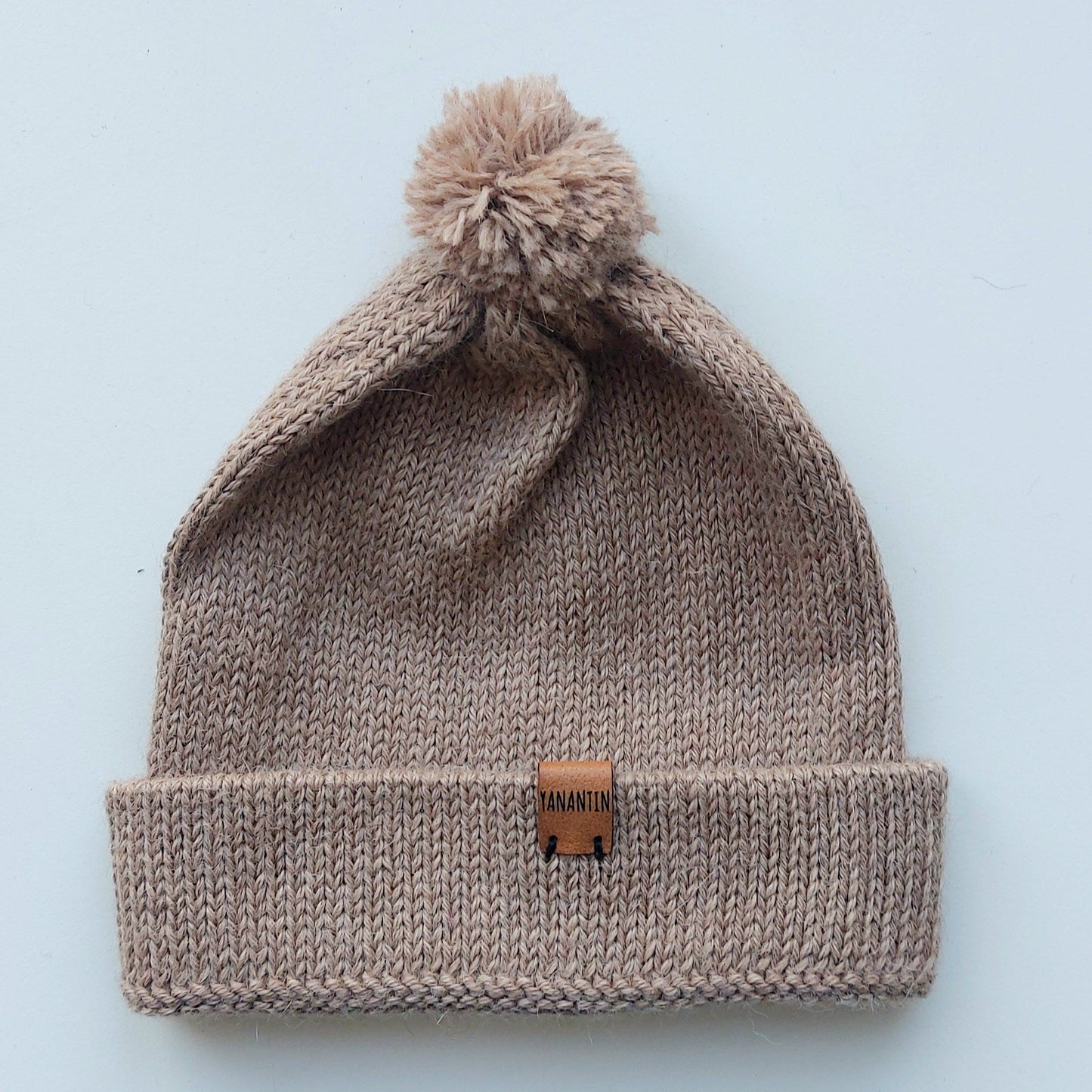 Light brown knitted baby hat with a fluffy pompom and a folded edge, handmade from 100% baby alpaca wool, featuring a soft vegan leather label.