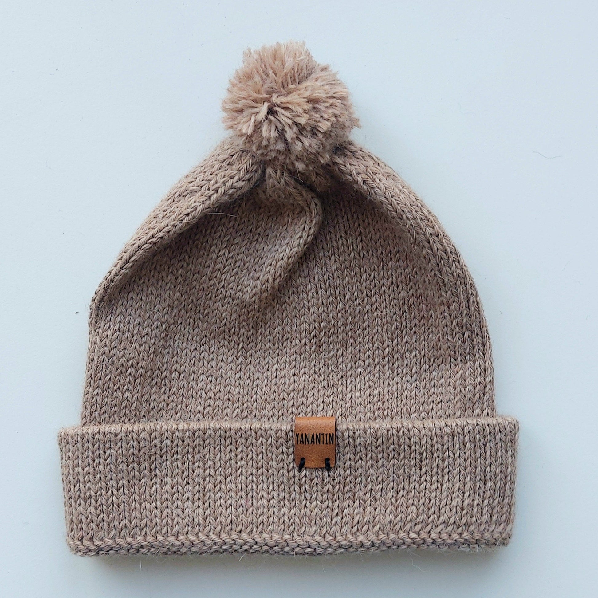 Light brown knitted baby hat with a fluffy pompom and a folded edge, handmade from 100% baby alpaca wool, featuring a soft vegan leather label.