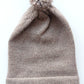 Light brown knitted baby hat with a fluffy pompom and a folded edge, handmade from 100% baby alpaca wool, featuring a soft vegan leather label.