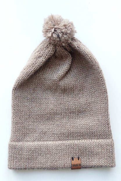 Light brown knitted baby hat with a fluffy pompom and a folded edge, handmade from 100% baby alpaca wool, featuring a soft vegan leather label.