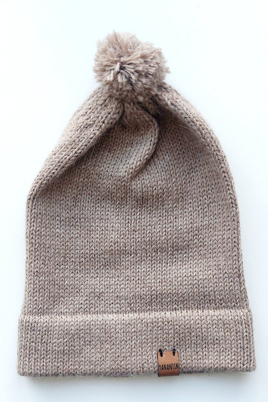 Light brown knitted baby hat with a fluffy pompom and a folded edge, handmade from 100% baby alpaca wool, featuring a soft vegan leather label.