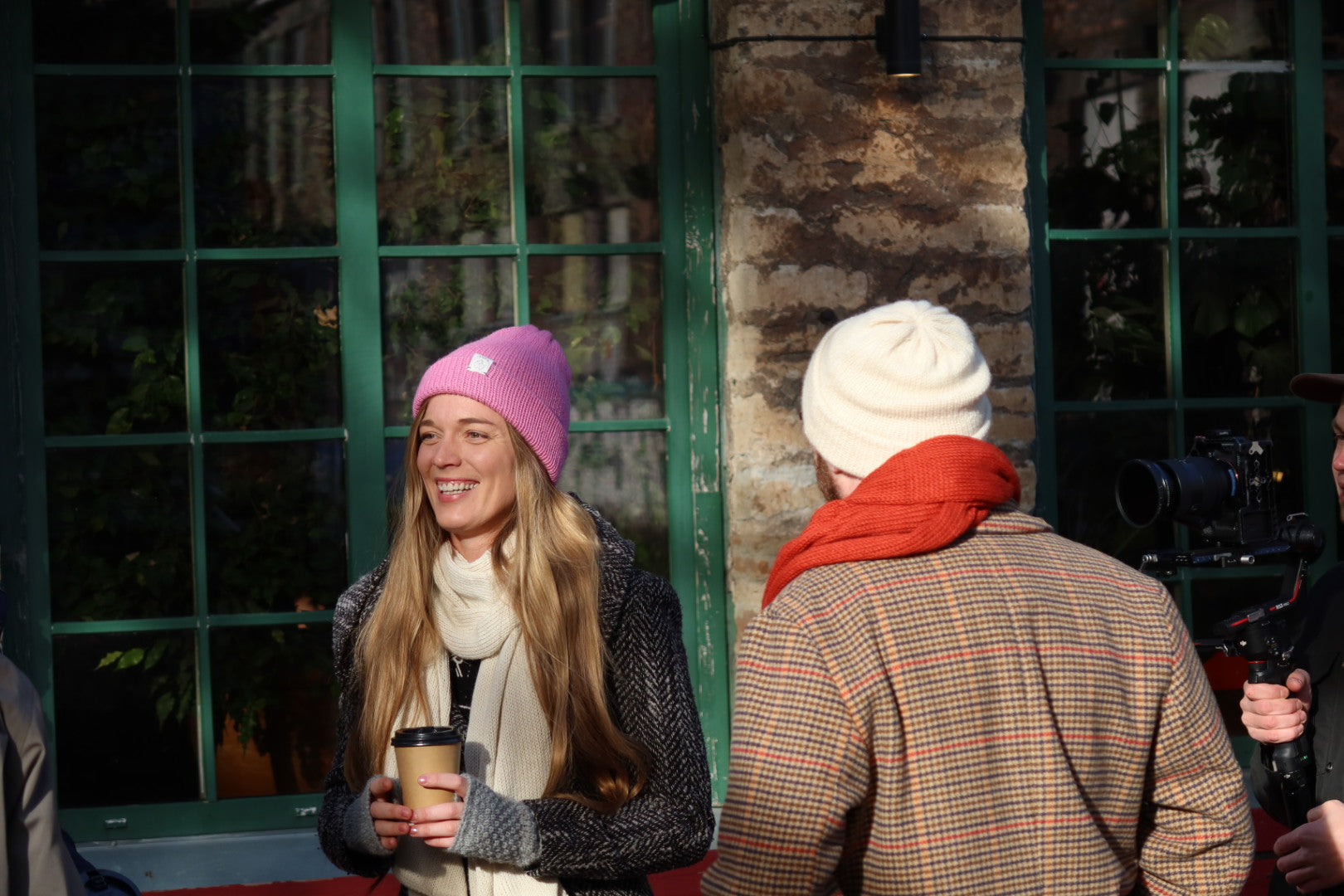 Two people wearing winter hats and scarves standing outside a building with a camera in the background.