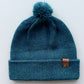Handmade peacock blue baby hat in soft 100% baby alpaca wool with a pompom, foldable edge, and brown vegan leather label, offering warmth and comfort.