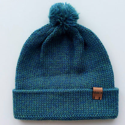 Handmade peacock blue baby hat in soft 100% baby alpaca wool with a pompom, foldable edge, and brown vegan leather label, offering warmth and comfort.
