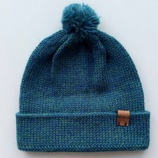 Handmade peacock blue baby hat in soft 100% baby alpaca wool with a pompom, foldable edge, and brown vegan leather label, offering warmth and comfort.