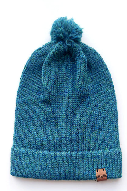 Handmade peacock blue baby hat in soft 100% baby alpaca wool with a pompom, foldable edge, and brown vegan leather label, offering warmth and comfort.